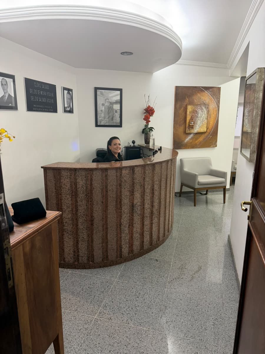 Recepcionista sorrindo atrás do balcão de atendimento da clínica, com os retratos na parede ao fundo